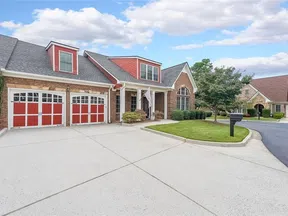 Suburban brick house with red accents, three-car garage, manicured lawn, and clear blue sky.