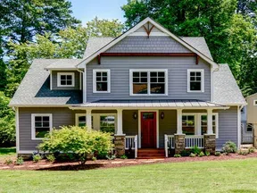 Two-story gray house with white trim, a red front door, stone pillars, and a manicured lawn in a wooded area.