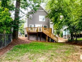 Backyard view of a gray two-story house with wooden deck and stairs surrounded by trees and grass.
