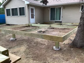 Wooden deck frame construction supported by concrete piers next to a beige house with windows and sliding door.