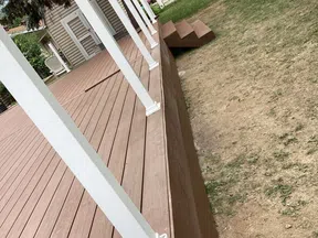 New brown wooden deck with white railing posts and small stairs leading to a backyard with patchy grass.