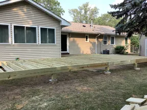 Wooden deck framework under construction beside a single-story house in a backyard with trees and grass.
