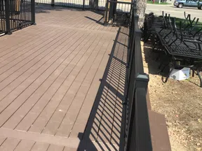 Empty brown wooden deck with black metal railing and stacked outdoor chairs beside trees in sunny weather.