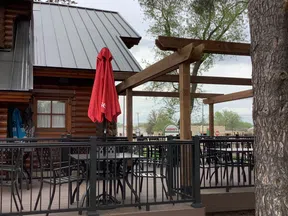 Outdoor seating area with black metal chairs and tables, red closed umbrella, wooden pergola, and log cabin exterior.