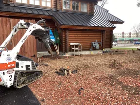 Mini excavator with auger attachment clearing rocky soil beside wooden cabin building under construction.
