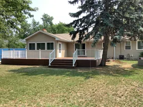 Single-story beige house with a large wooden deck, white railing, and green lawn under a tall tree.