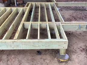 Wooden deck frame construction with metal supports next to a log cabin on a dirt ground.