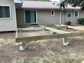 Wooden deck foundation frame built on concrete piers next to a beige house with sliding glass door.