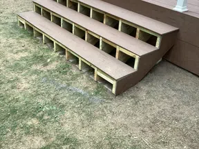Three-step brown wooden outdoor stairs attached to a deck with visible support structure on dry lawn.