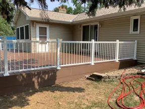 Raised brown wooden deck with white railings attached to beige house under tree shade in backyard.