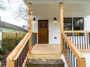 Front porch of a house with wooden railings, a wooden door, white siding, and outdoor wall lights.