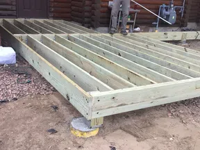 Construction worker building wooden deck frame outside a log cabin on a dirt and gravel surface.