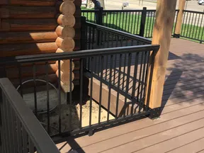 Brown wooden deck with black metal railing and part of a log cabin under clear daylight.