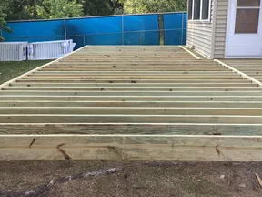 Construction of a wooden deck frame attached to a house in a backyard with trees and a blue fence.