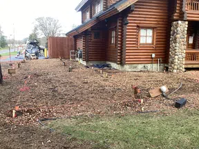 Prepared ground with stakes and markings next to a log cabin under construction with stone pillar details.