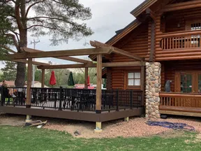 Outdoor wooden deck with black railing and pergola attached to a rustic log cabin surrounded by green grass.