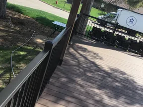 Spacious backyard deck with brown wooden flooring, black metal railing, and a white truck parked nearby on a sunny day.