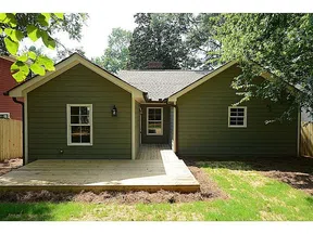 Green single-story house with white trim, wooden deck, and lush backyard surrounded by trees and fence