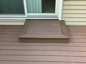 Brown wooden deck with a single step leading up to sliding glass door and light beige siding on house.