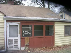 Old house exterior with damaged screen door and boarded-up wooden panel beneath three windows.