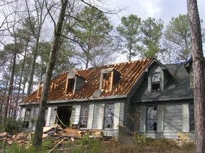 Old gray house with roof stripped down to wooden rafters surrounded by trees and debris on the ground