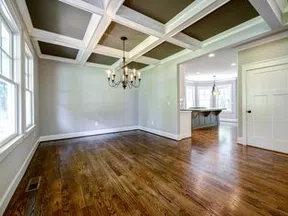Empty room with hardwood floors, large windows, coffered ceiling, and chandelier lighting in a modern house.