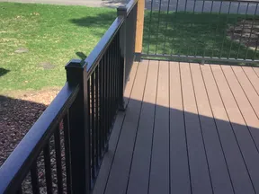 Outdoor deck with dark brown wooden floorboards and black metal railing overlooking a road and green lawn.