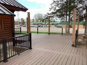 Spacious wooden deck with black metal railings and vertical posts next to a log cabin under a blue sky.