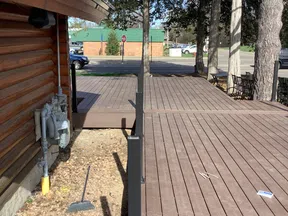 Wooden deck adjacent to a log cabin with metal railing, trees, broom, and parked cars in the background.
