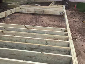 Wooden deck frame under construction on dirt ground with nearby trees and road in the background.