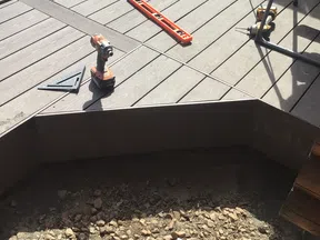 Wooden deck under construction with power drill, level, and rocky ground below the deck frame.
