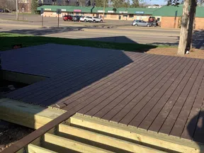 Partially completed wooden deck with exposed beams in foreground and street with buildings and parked cars in background.