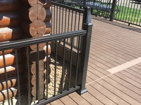 Brown wooden deck with black metal railing attached to a log cabin corner, outdoor suburban setting.