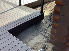 Close-up of brown composite decking and railing next to a log cabin corner with exposed ground and cables.
