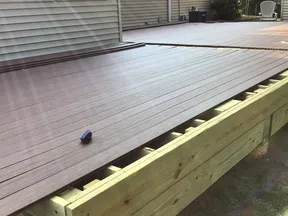 Partially installed dark composite decking boards on a wooden deck frame next to a house exterior in daylight.
