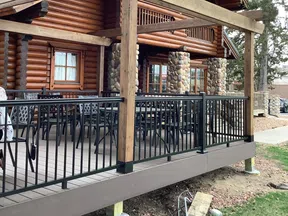 Outdoor deck with black metal railing attached to a log cabin featuring stone pillars and wooden beams.