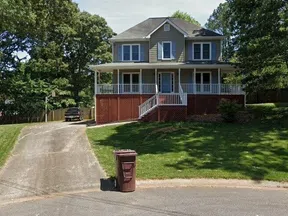 Two-story house with gray siding, white porch railing, red brick foundation, driveway, and green lawn on a sunny day.