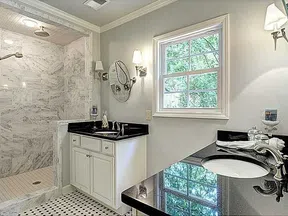 Modern bathroom with marble shower, dual sinks with black countertops, white cabinetry, and natural light from window.