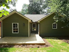 Green single-story house with wooden deck, white trim windows, and surrounding trees in a backyard.