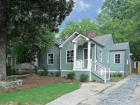 Charming single-story gray house with white trim, front porch, greenery, and a driveway leading to a backyard gate.