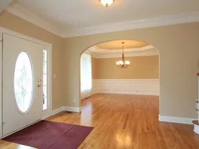 Bright empty living room with hardwood floors, beige walls, white trim, oval glass front door, and chandelier.