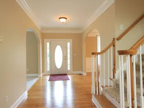 Bright entryway with wooden floors, staircase with carpeted steps, beige walls, and white door with oval glass window.