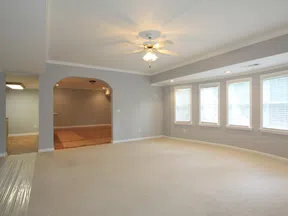 Bright empty living room with beige carpet, gray walls, ceiling fan, and three large windows with blinds.