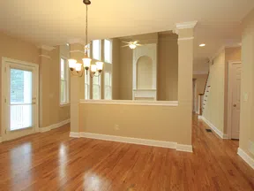 Empty spacious room with hardwood floors, beige walls, chandelier, ceiling fan, and large windows letting in natural light