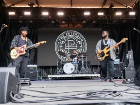 Three musicians performing on stage with guitars and drums, with The Peterson logo in the background
