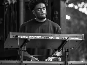 Black and white photo of a musician playing dual keyboards on stage with focused expression.