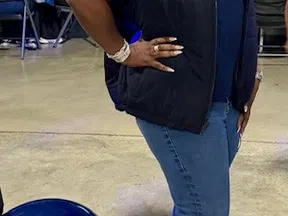 Smiling woman in blue vest and jeans posing indoors near a blue balloon with people seated in background.