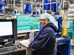 Smiling factory worker wearing hairnet and glasses works at computer station surrounded by industrial equipment.