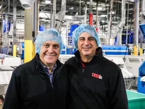 Two men wearing blue hairnets and black jackets smiling inside a large industrial manufacturing facility.