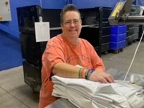 Smiling worker in orange shirt folding white linens on metal table in industrial laundry facility.
